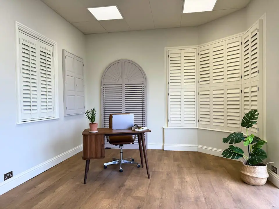 small room with desk and spinning chair, wooden flooring, green plant in the corner and small white shutters